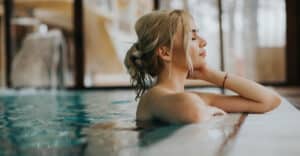 Femme blonde détendue, yeux clos, bras sur le bord d'une piscine de spa. Des gouttelettes d'eau perlent sur sa peau. Ambiance calme.