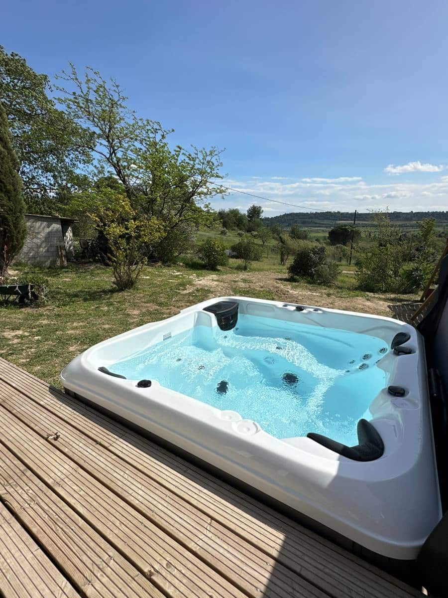 Jacuzzi extérieur bouillonnant sur une terrasse en bois, avec un paysage verdoyant et un ciel bleu dégagé en arrière-plan.