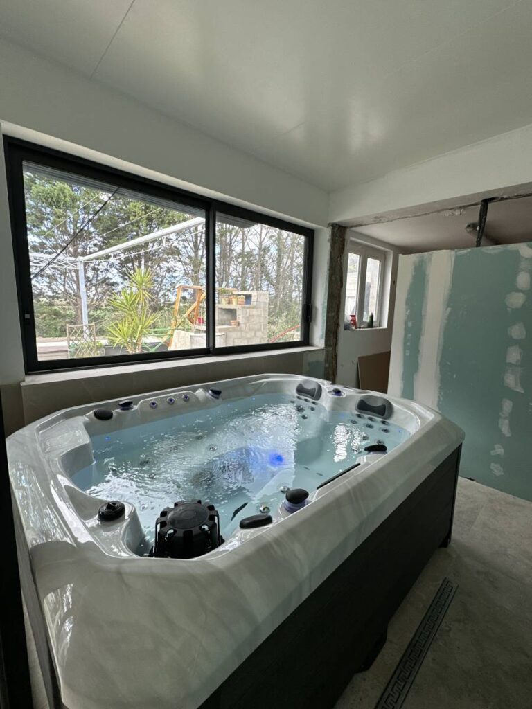 Nouveau jacuzzi bleu lumineux en fonctionnement dans une pièce en rénovation, face à une grande fenêtre donnant sur un jardin verdoyant. Mur en placo et sol brut.