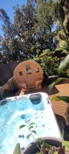 Jacuzzi bouillonnant sur une terrasse en bois, avec sauna tonneau et verdure sous ciel bleu. Espace détente et bien-être extérieur.