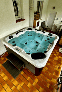 Jacuzzi moderne avec eau bleue lumineuse et bulles, éclairage LED, marches sombres, sur carrelage terre cuite sous une fenêtre avec barreaux.