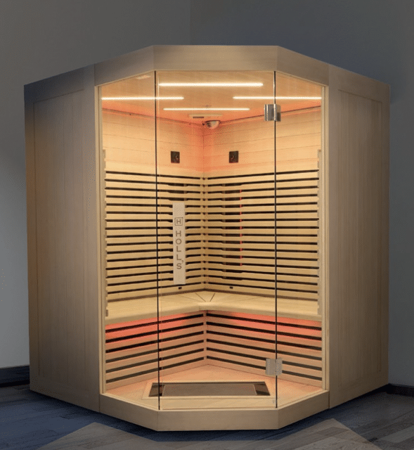 Sauna Infrarouge HOLLS : Design Moderne et Bien-être Sauna d'angle moderne en bois clair avec porte vitrée, éclairage LED blanc au plafond et rouge sous les bancs. Le panneau vertical à l'intérieur affiche le texte 'HOLLS'.