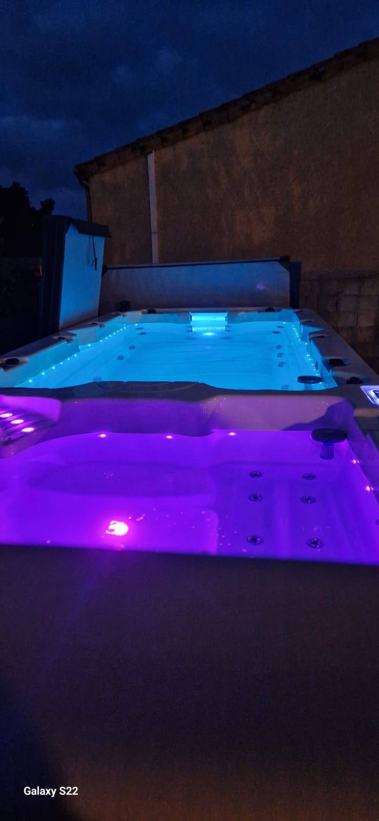Grand spa extérieur éclairé en bleu et violet à la tombée de la nuit, avec un mur de maison en arrière-plan. Galaxy S22.