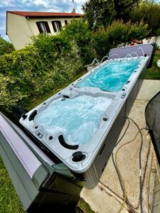 Spa de nage extérieur avec jacuzzi bouillonnant et jets d'eau, installé près d'une maison avec toit rouge et végétation luxuriante.