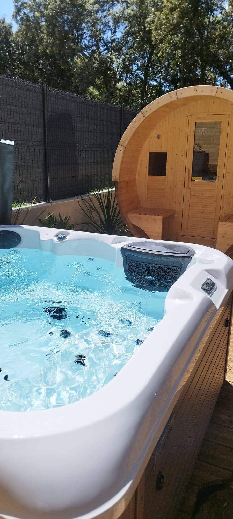 Spa extérieur moderne avec eau bouillonnante, juxtaposé à un sauna baril en bois HOLL'S. Clôture et verdure au soleil.