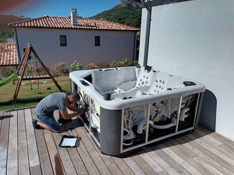 Réparation Jacuzzi & Spa : Dépannage par Technicien Technicien agenouillé réparant un jacuzzi sur une terrasse en bois. Les panneaux du spa sont ouverts, révélant les tuyaux et pompes.