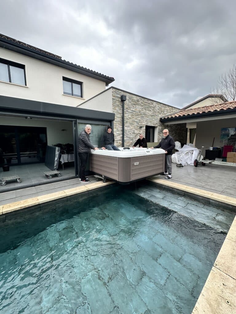 Installation spa Yendon : le jacuzzi parfait pour la piscine Quatre hommes installent un jacuzzi Yendon marron sur roulettes, le positionnant au bord d'une piscine moderne dans un jardin extérieur.