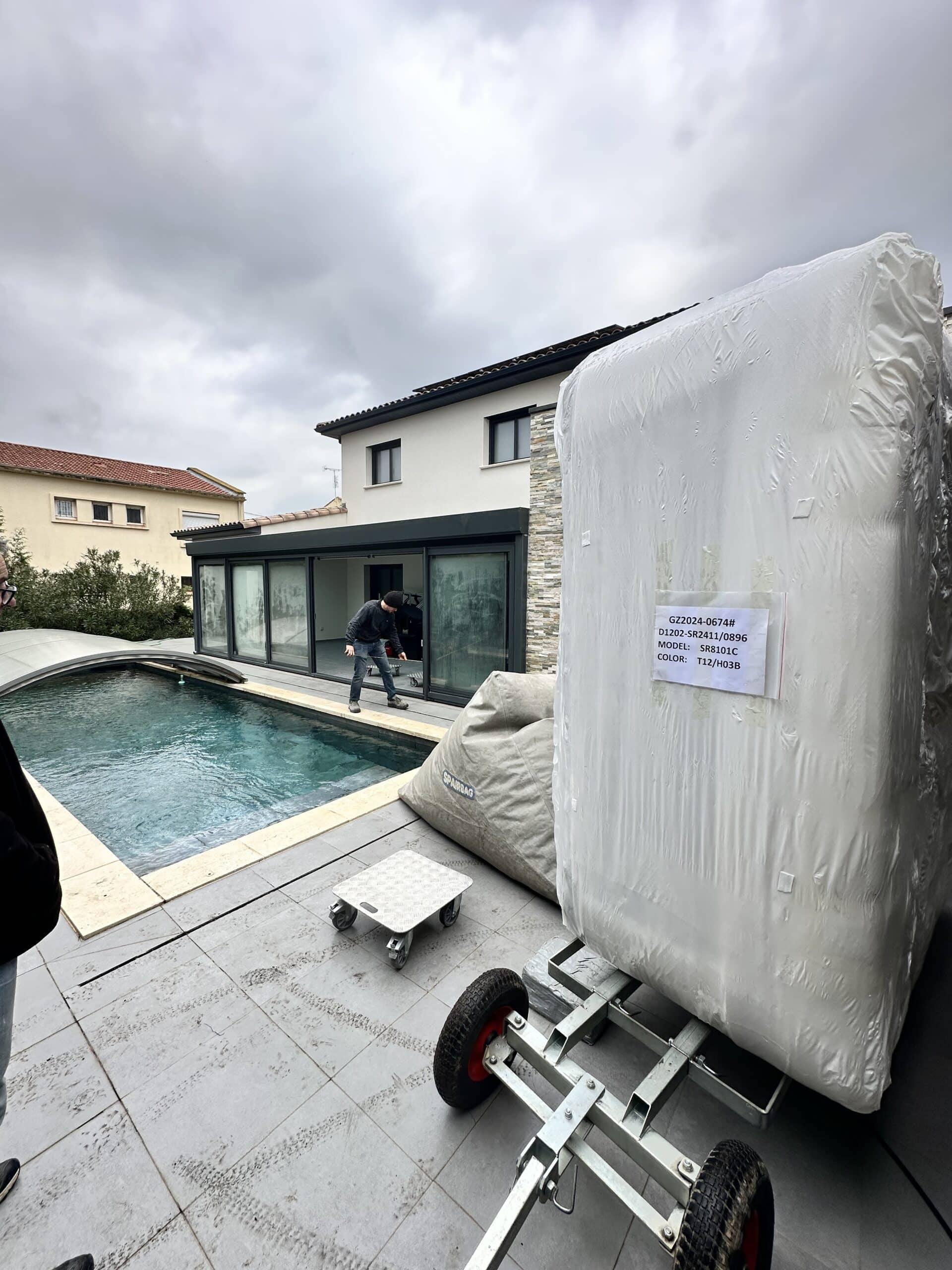 Grosse pompe à chaleur SR8101C enveloppée sur un chariot de livraison devant une piscine et une maison moderne. L'installateur vérifie l'accès. Ciel gris.