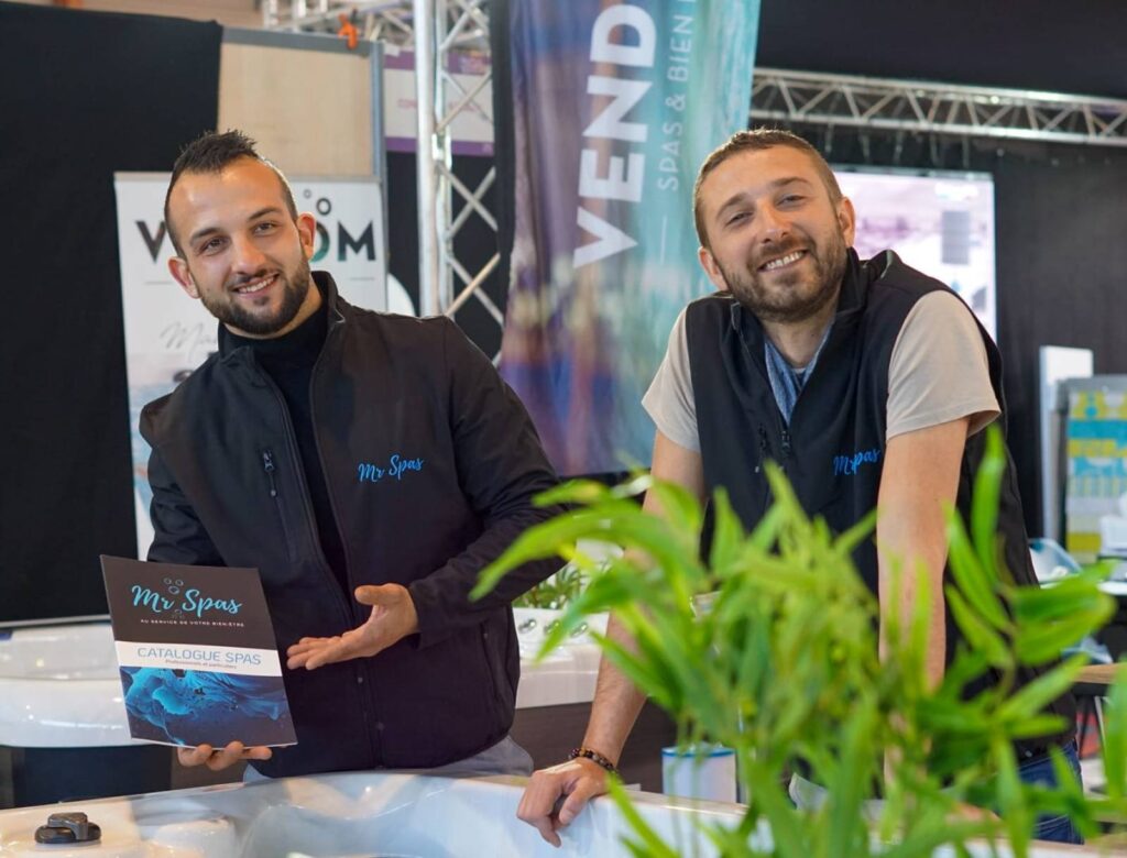 Mr Spas : Catalogue et Experts en Spas Professionnels Deux hommes souriants de Mr Spas présentent un catalogue de spas lors d'un événement, avec une affiche 'VEND' en arrière-plan.