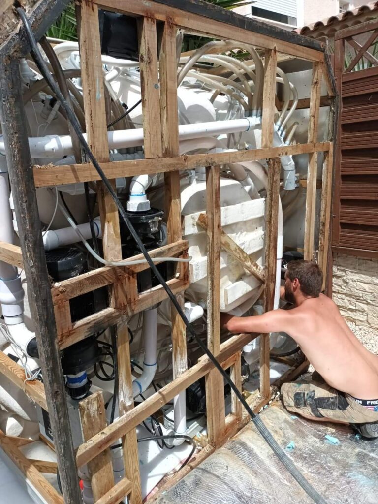 Réparation spa jacuzzi : diagnostic et entretien de la plomberie. Technicien répare la plomberie complexe (pompes, tuyaux PVC) d'un spa/jacuzzi exposé. Structure en bois brute visible.