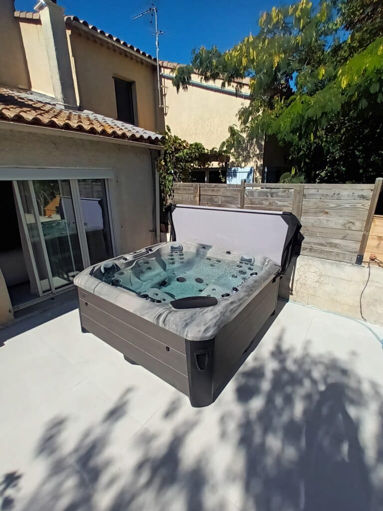 Spa extérieur : Détente sur terrasse carrelée Spa extérieur avec couverture ouverte, bulles visibles dans l'eau, installé sur une terrasse carrelée claire, sous un ciel bleu et ombragé par des arbres.