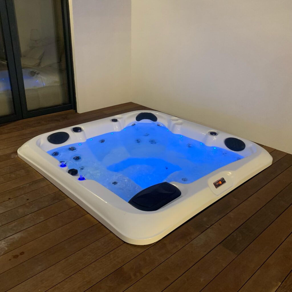 Spa gonflable carré blanc éclairé par des LED bleues, en activité sur une terrasse en bois sombre près d'une baie vitrée.