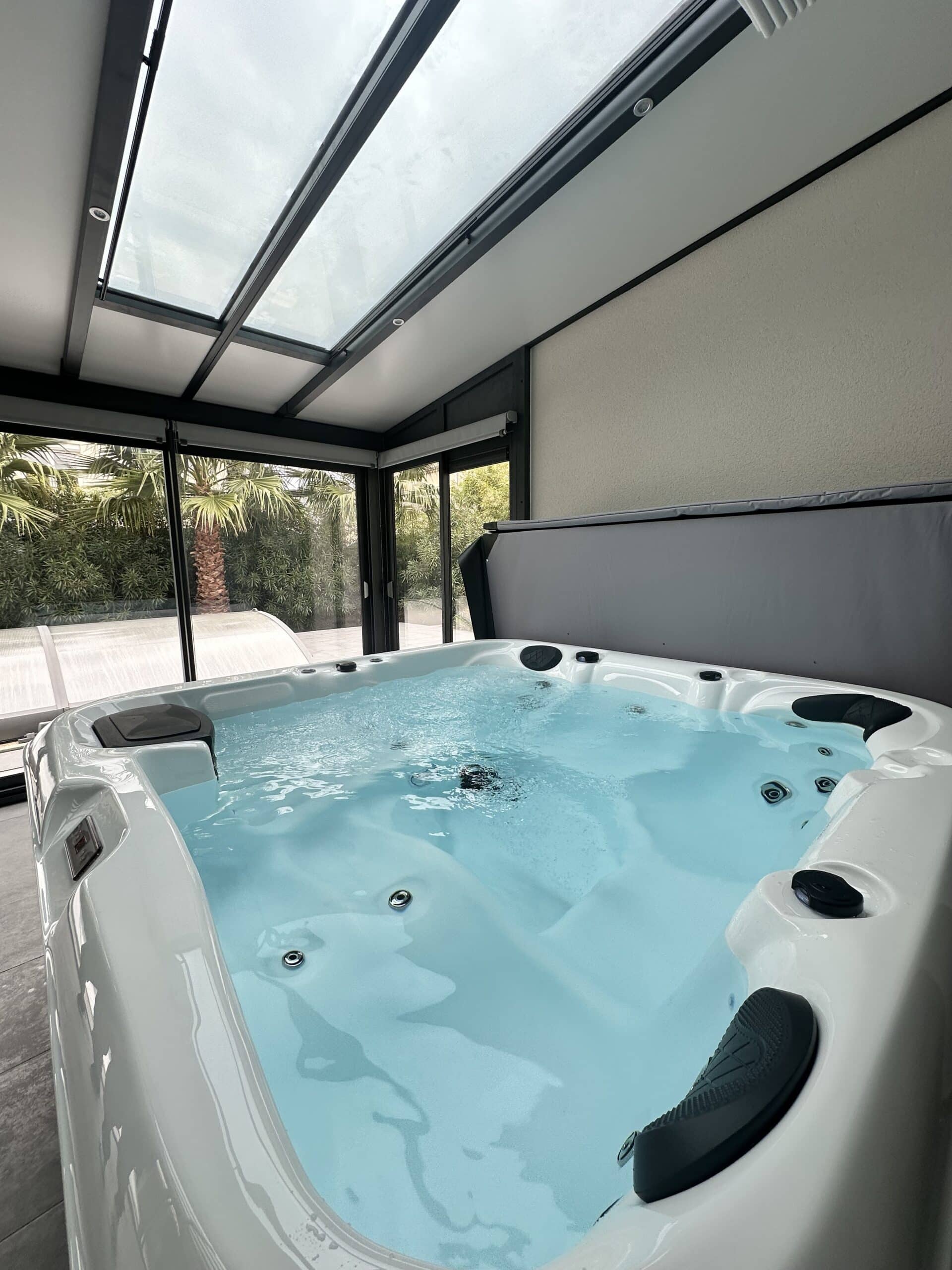 Spa blanc moderne en véranda vitrée. L'eau turquoise et bouillonnante reflète la lumière du toit en verre. Vue extérieure sur la végétation et palmiers.