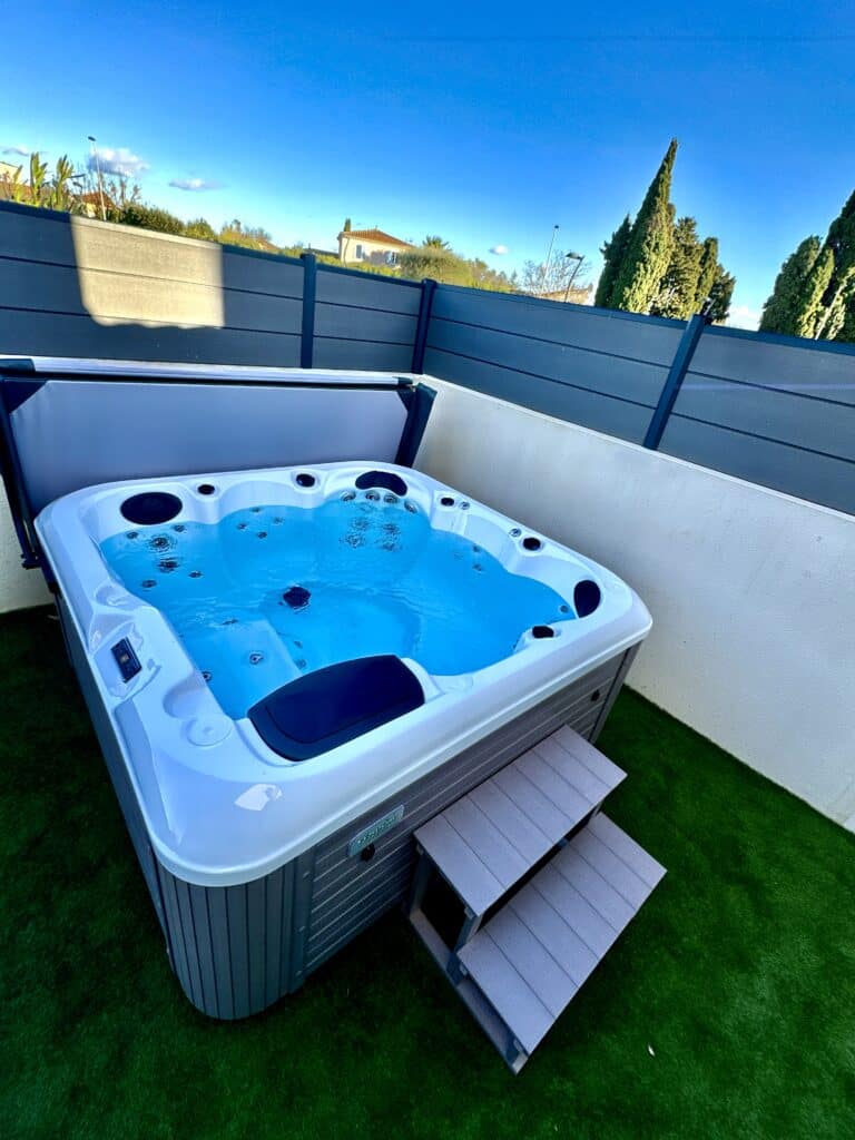 Spa extérieur moderne Vendôm à l'eau bleue bouillonnante, sur gazon synthétique. Installation en terrasse privée avec clôture gris foncé.