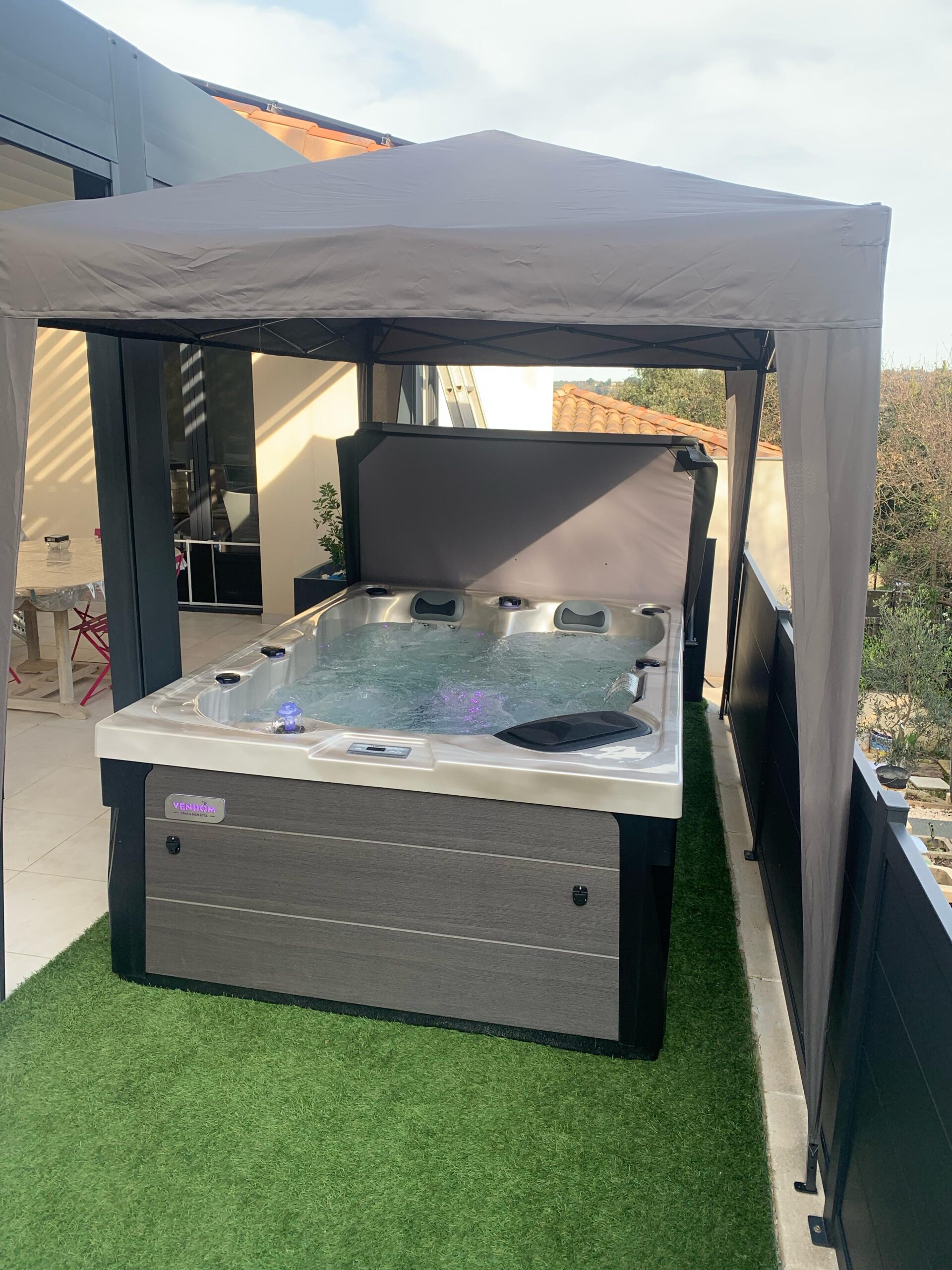 Spa Jacuzzi VENIDOM extérieur, eau bouillonnante éclairée sous un auvent gris de protection, sur gazon synthétique.