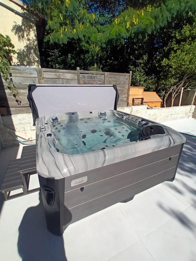 Spa extérieur Vendom avec couverture ouverte, rempli d'eau bouillonnante, installé sur une terrasse blanche en plein soleil.