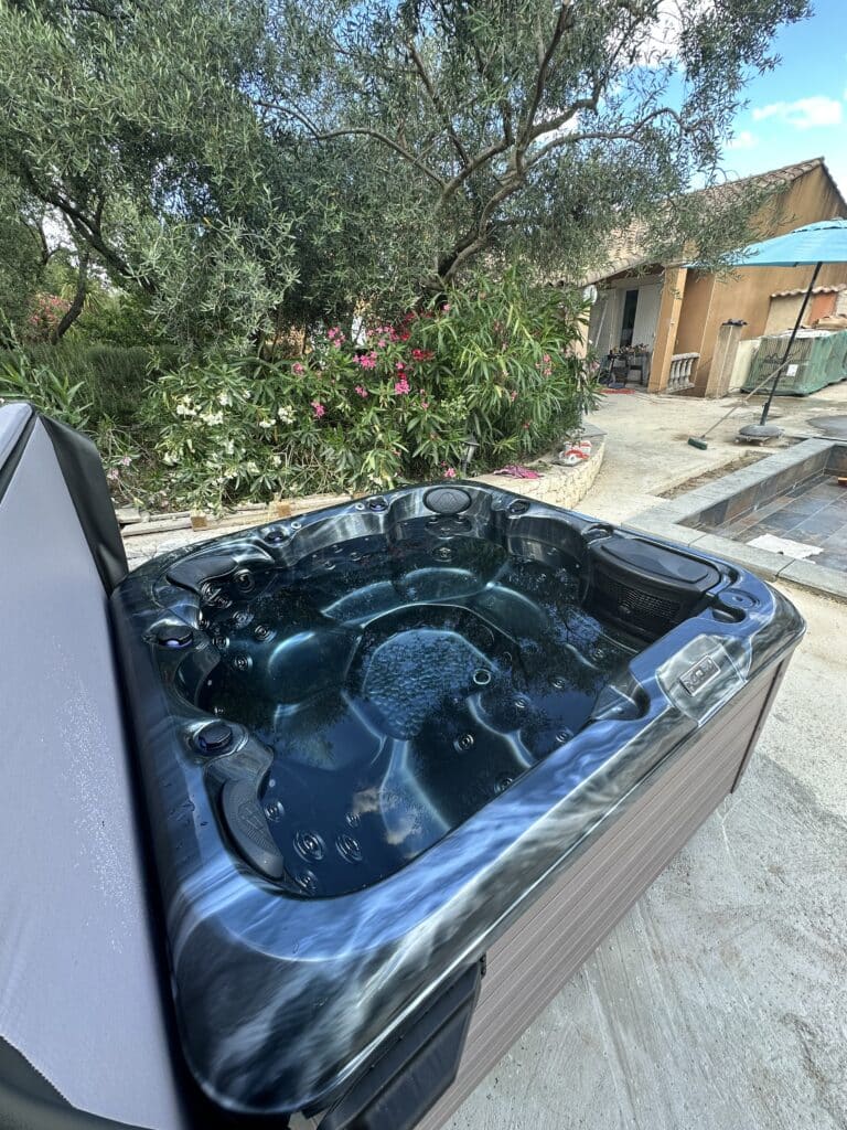 Spa Jacuzzi extérieur : Installation sous un olivier. Spa jacuzzi d'extérieur bleu-gris neuf, installé sur une terrasse en béton ensoleillée, sous un olivier. Fleurs roses et maison en arrière-plan.
