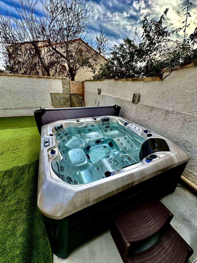Spa Jacuzzi extérieur : profitez d'une détente optimale. Spa extérieur moderne avec eau turquoise bouillonnante sur terrasse. Murs blancs, gazon synthétique et ciel bleu dramatique en arrière-plan.