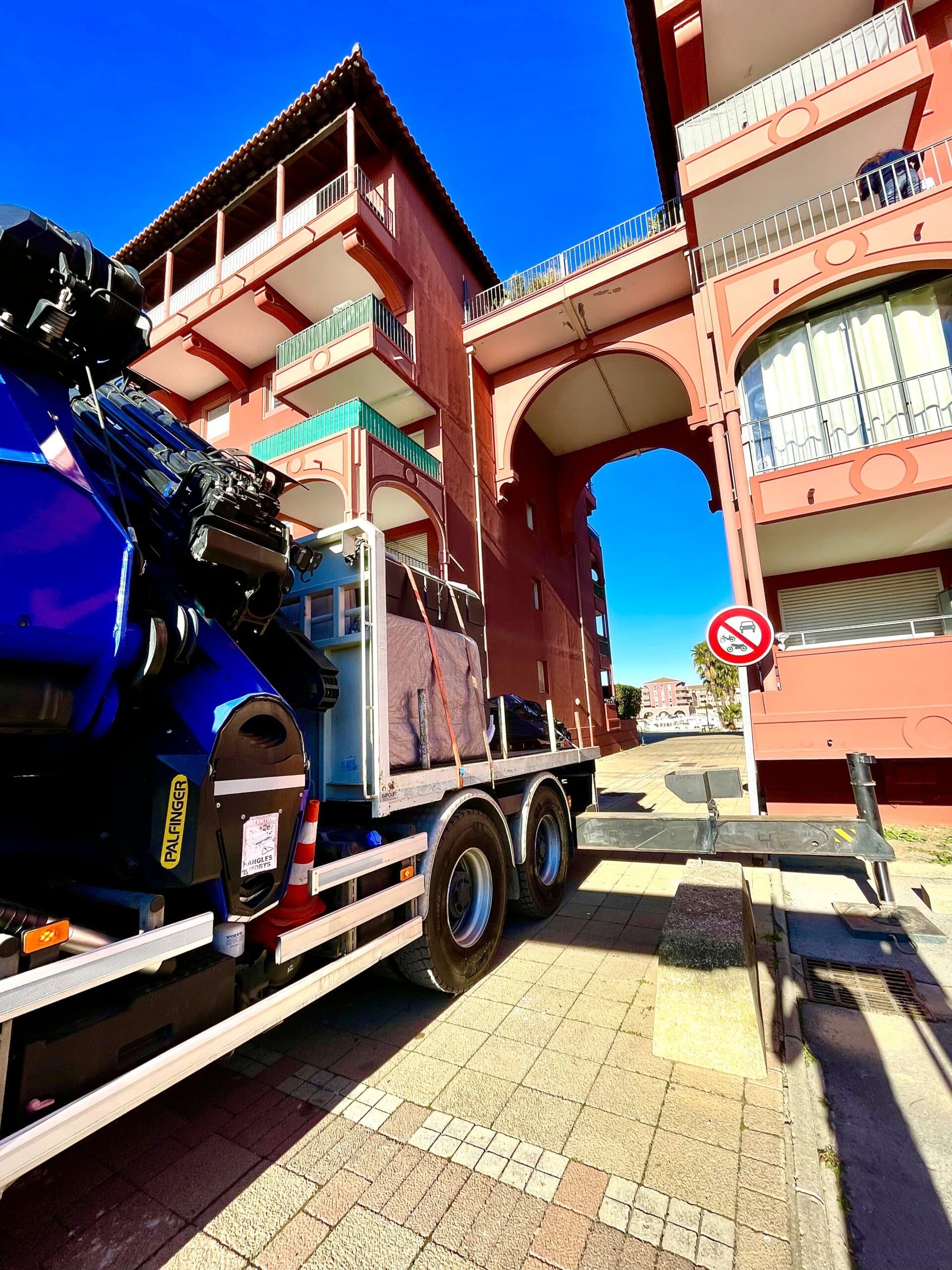 Camion grue Palfinger bleu garé près d'un immeuble en terre cuite avec balcons et arches. Panneau Angles Morts et interdiction de circuler.