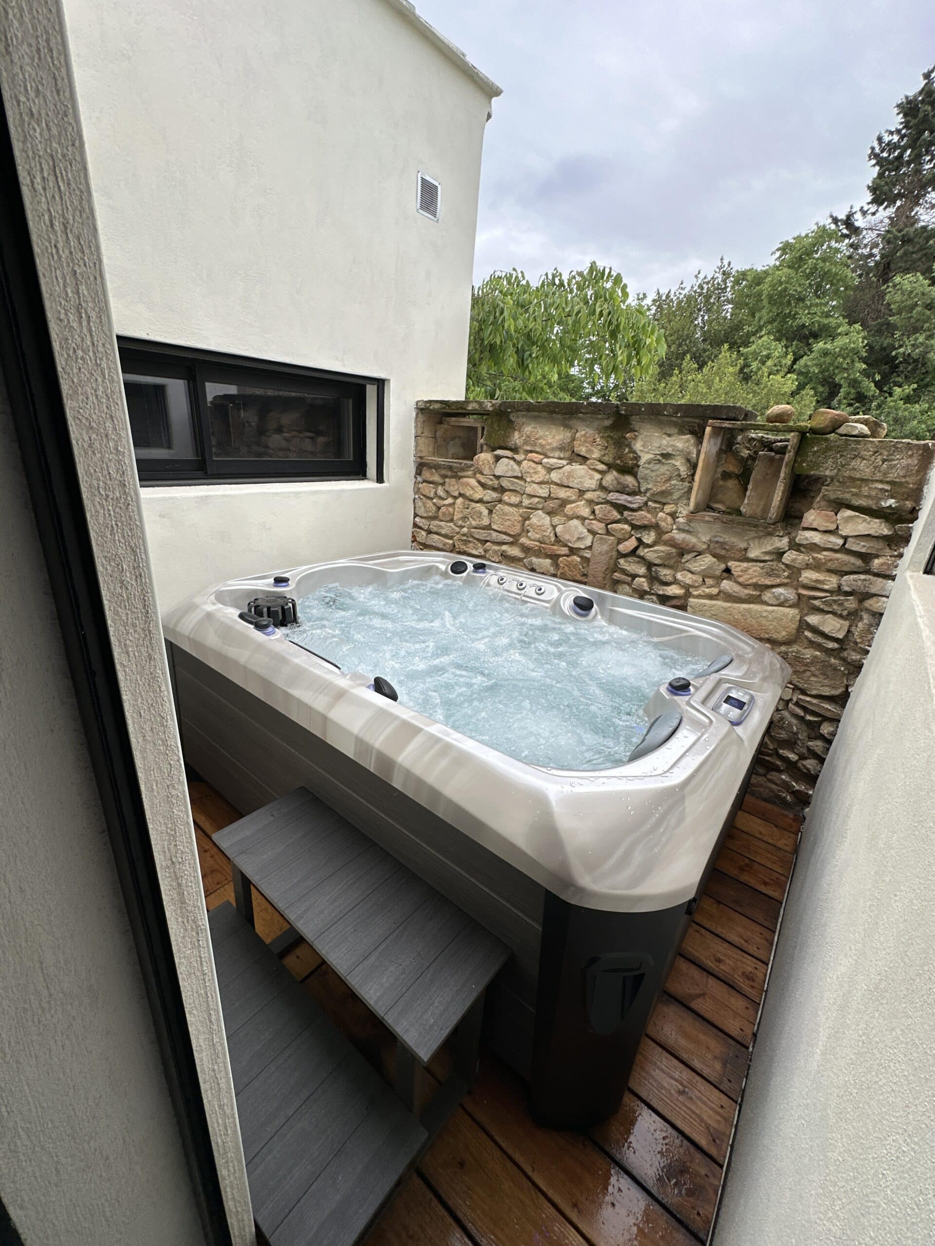 Grand bain à remous extérieur bouillonnant sur une terrasse en bois. Espace privé entre murs blancs et mur de pierre sèche ancien.