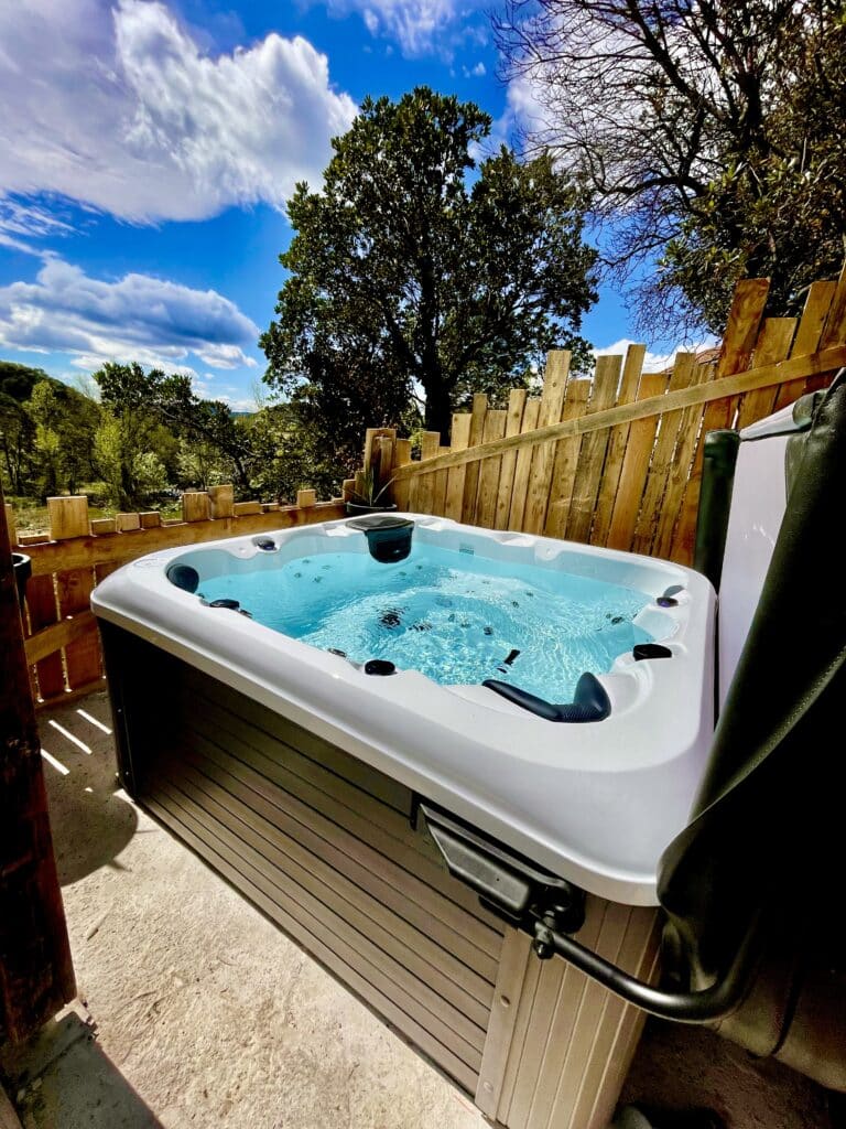 Hot Tub Extérieur: Luxe et Détente au cœur de la Nature. Grand jacuzzi extérieur avec eau bleue, installé près d'une palissade en bois. Vue imprenable sur une forêt dense et des collines sous un ciel bleu.