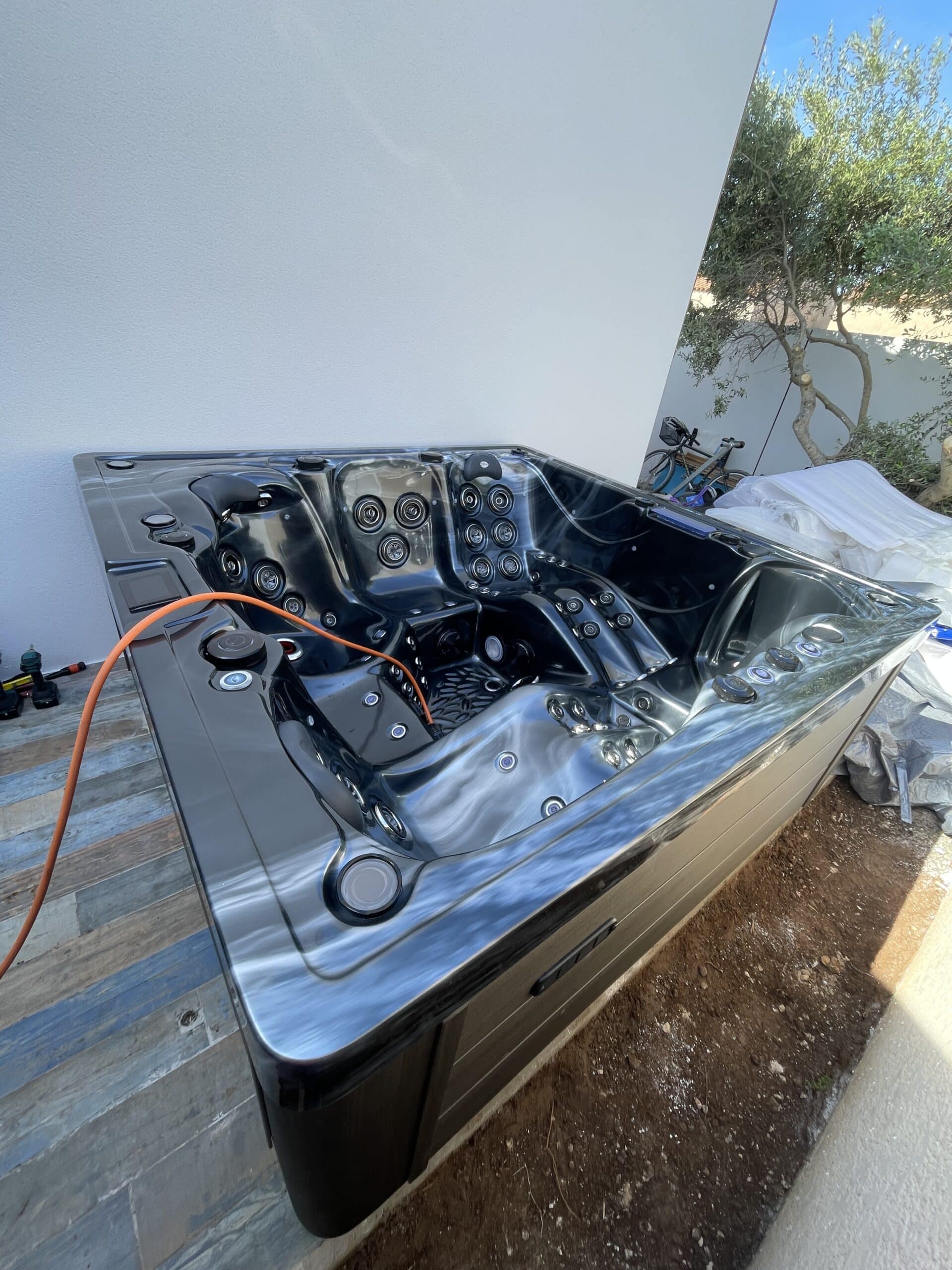 Spa jacuzzi extérieur noir moderne avec intérieur brillant, en cours d'installation contre un mur blanc près d'un olivier.