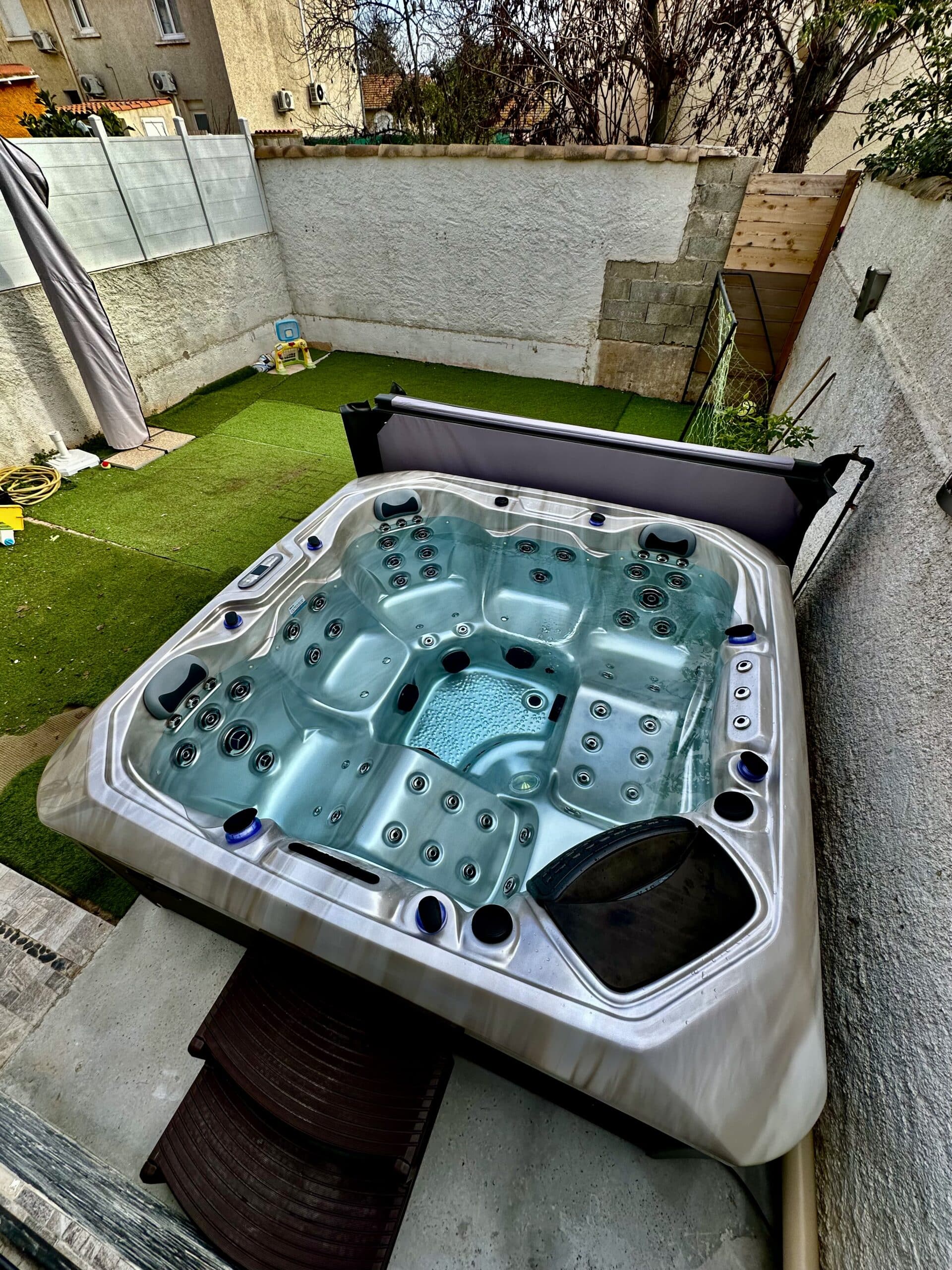 Grand jacuzzi extérieur moderne, eau bleue scintillante, installé sur une terrasse avec gazon synthétique et murs d'enceinte.