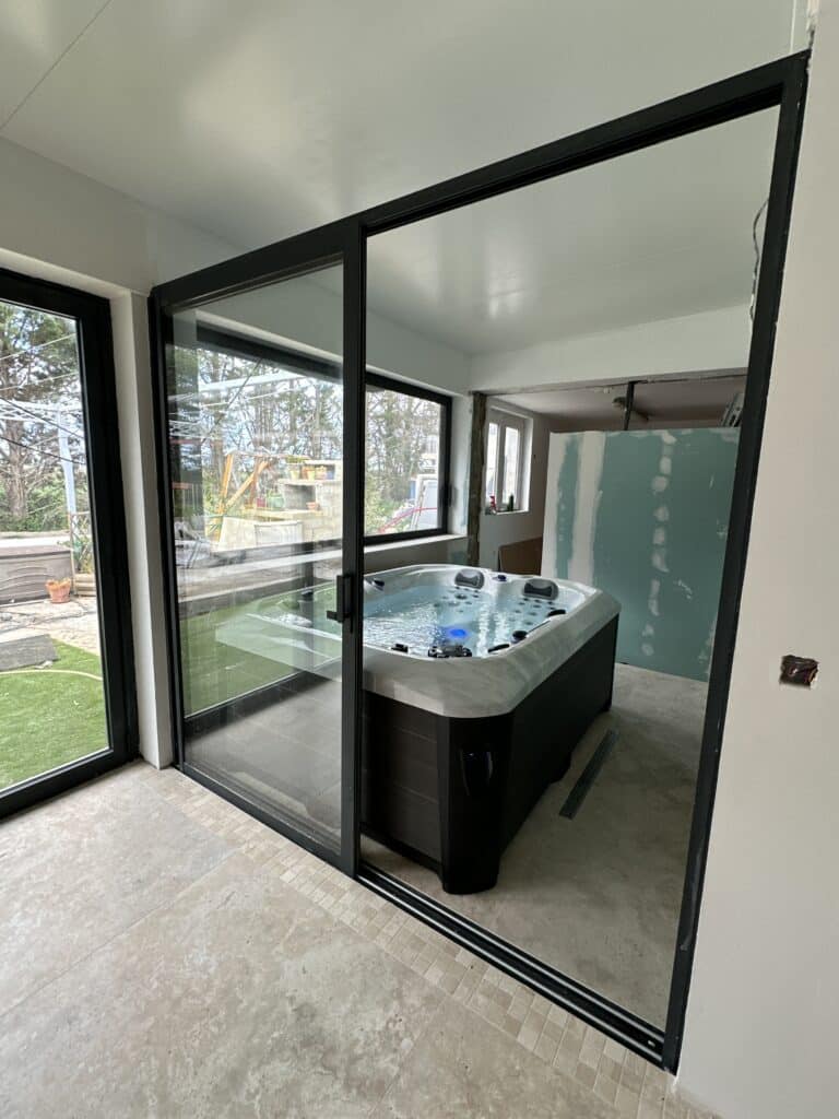 Installation spa intérieur : portes vitrées et rénovation Jacuzzi moderne installé dans une pièce en rénovation, séparé par de grandes portes coulissantes vitrées noires. Sol en travertin.