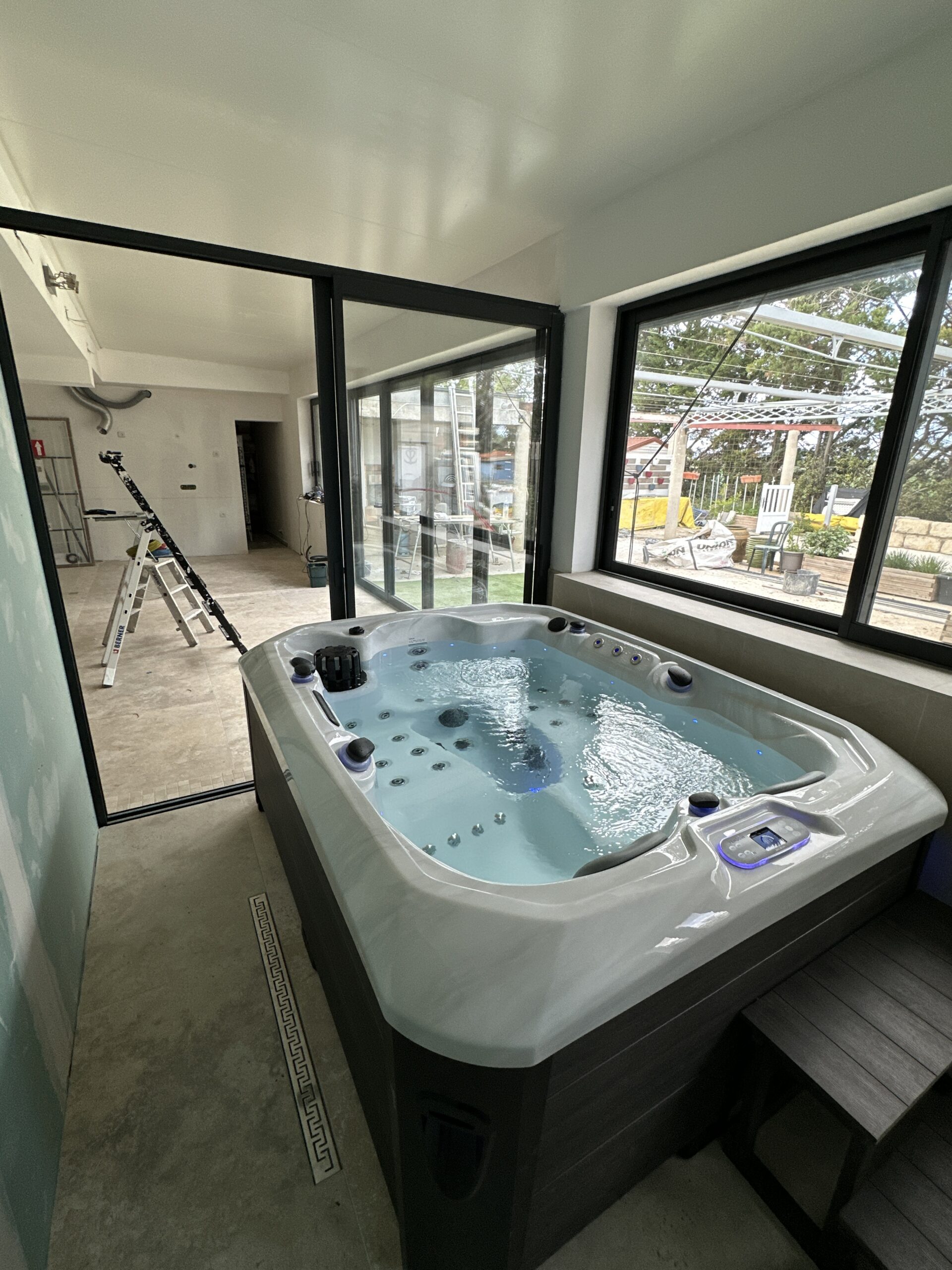 Spa jacuzzi intérieur en fonction, installé dans une pièce en construction. Grandes baies vitrées noires et drain au sol.