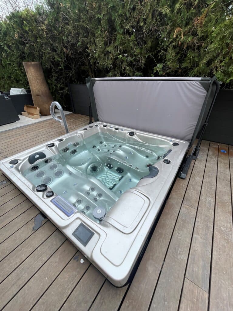 Spa de luxe sur terrasse en bois | Couverture relevée Spa de luxe moderne installé sur une terrasse en bois mouillée. La couverture est relevée par un lève-couverture. Haie verte en arrière-plan.