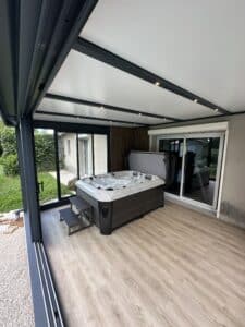 Véranda moderne avec sol imitation bois et plafond blanc, abritant un grand spa jacuzzi en bulles. Structure en aluminium anthracite ouverte sur le jardin.