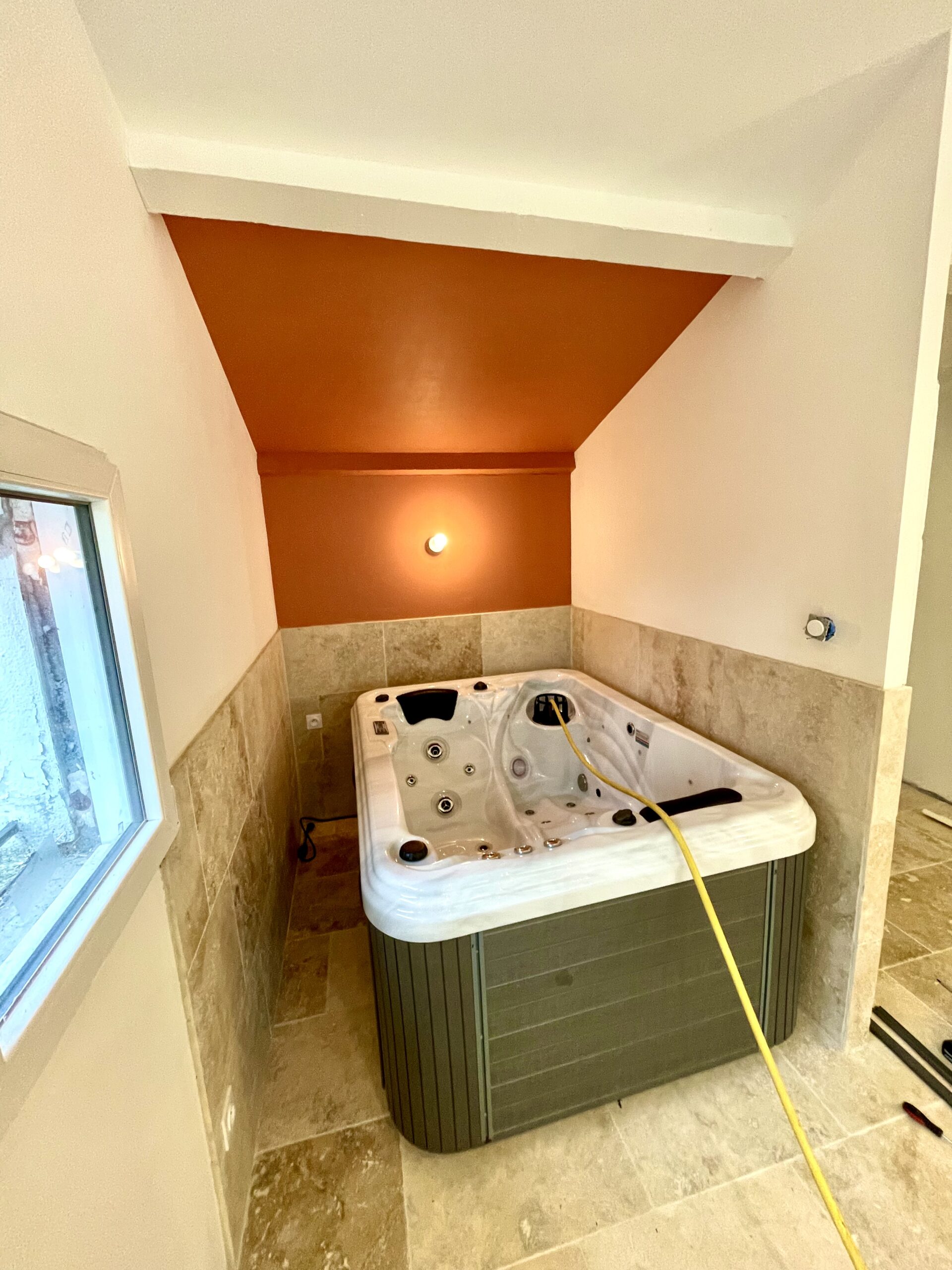 Spa blanc et gris installé dans une alcôve. Murs en carrelage travertin et plafond orange vif éclairé par un spot. Ambiance chaleureuse.