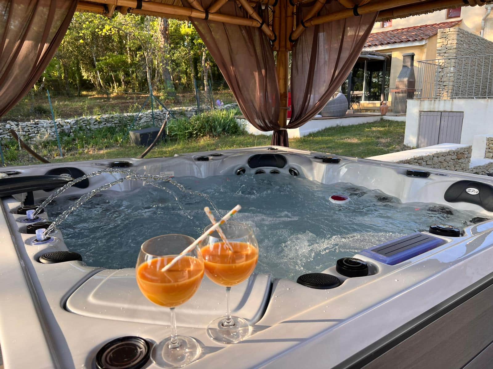 Jacuzzi extérieur en marche sous pergola, jets d'eau actifs. Deux cocktails sur le rebord. Cadre de détente près d'une villa en pierre et la forêt.