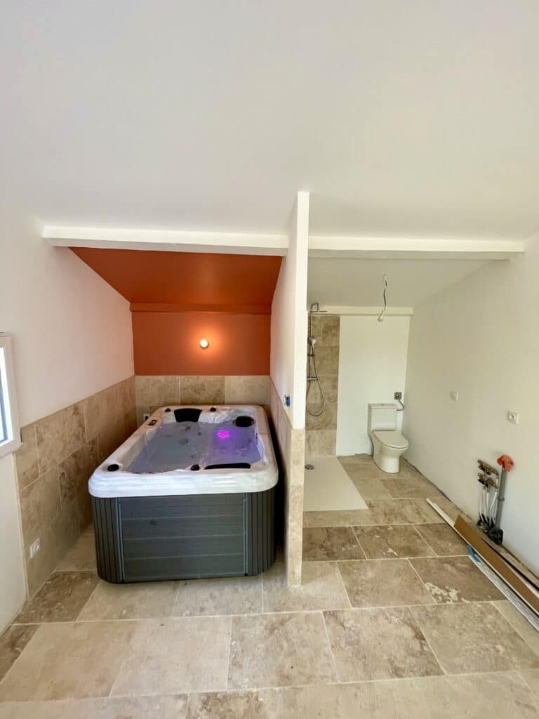 Jacuzzi, douche et WC : salle de bain moderne en travertin. Salle de bain/spa en rénovation. Jacuzzi sous plafond orange vif, douche et WC. Carrelage en travertin clair murs et sol.