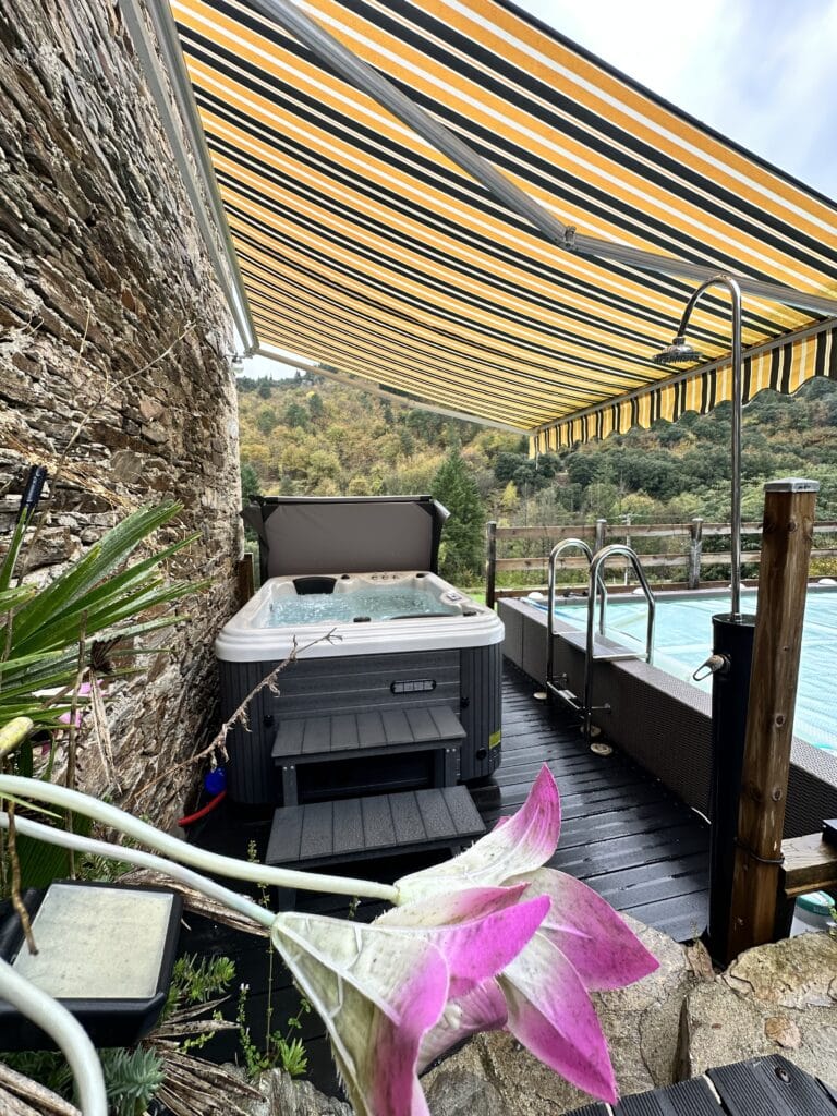 Jacuzzi extérieur et piscine sous un grand auvent rayé jaune et noir. Terrasse en bois sombre près d'un mur de pierre rustique face aux collines boisées.