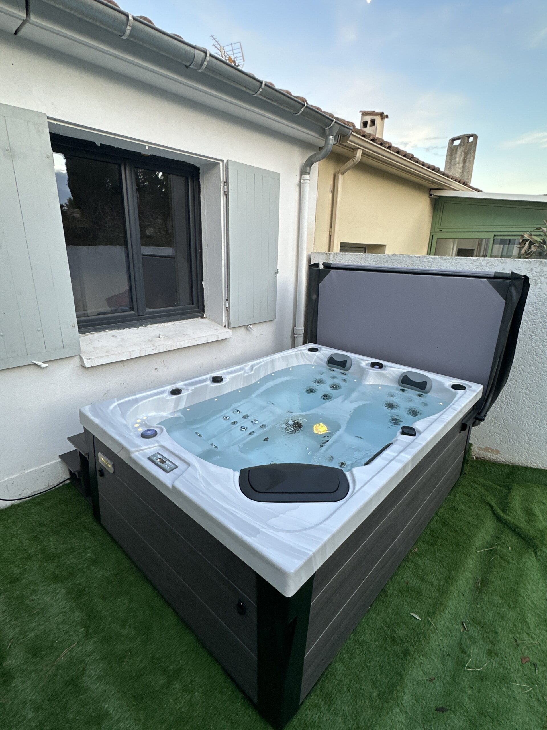 Jacuzzi extérieur moderne, coque blanche et armoire grise, installé sur gazon synthétique. Le spa bouillonne, contre une façade de maison aux volets gris.