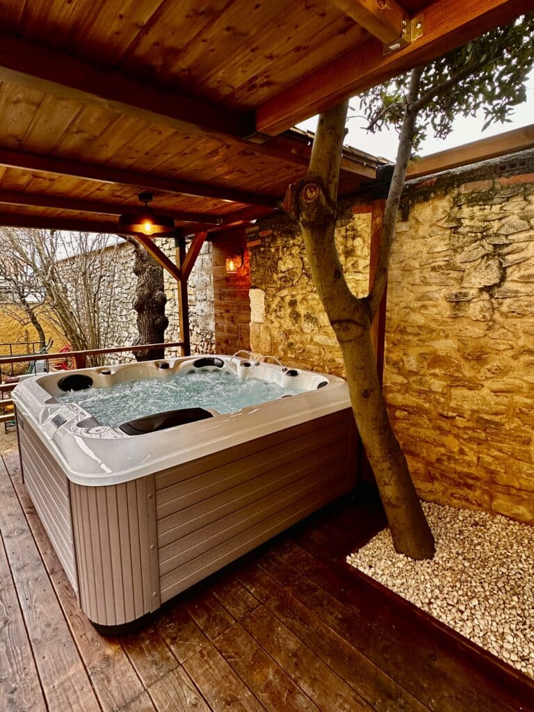 Jacuzzi extérieur couvert : ambiance bois et murs en pierre Spa jacuzzi extérieur bouillonnant sous pergola en bois. L'espace est entouré de murs en pierre rustiques et éclairé par des lumières chaudes.