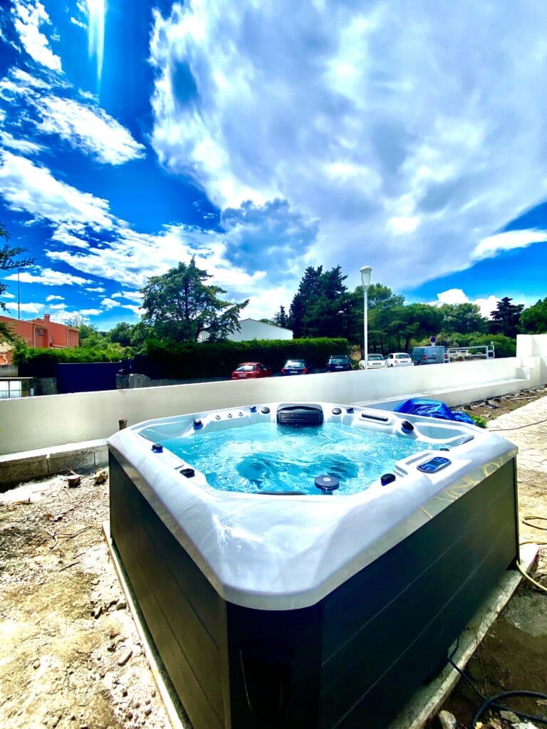 Jacuzzi extérieur : installation sous un ciel bleu éclatant Jacuzzi moderne extérieur bleu vibrant sur un chantier, encadré de verdure et d'un mur blanc, sous un ciel bleu intense et nuageux.