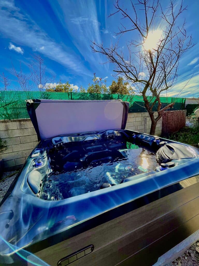 Jacuzzi extérieur ouvert en journée ensoleillée. La cuve bleu profond reflète le ciel intense et les rayons du soleil.