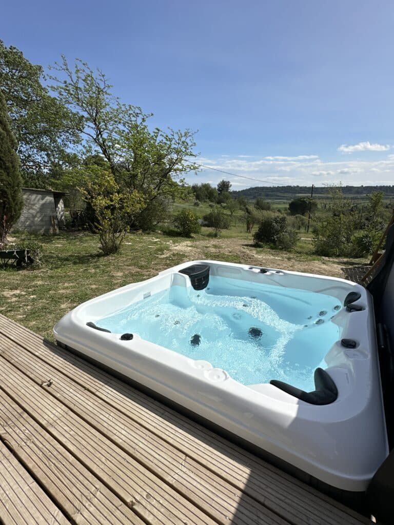 Spa jacuzzi blanc extérieur en fonction sur une terrasse en bois, offrant une vue dégagée sur la nature verdoyante sous un ciel bleu.