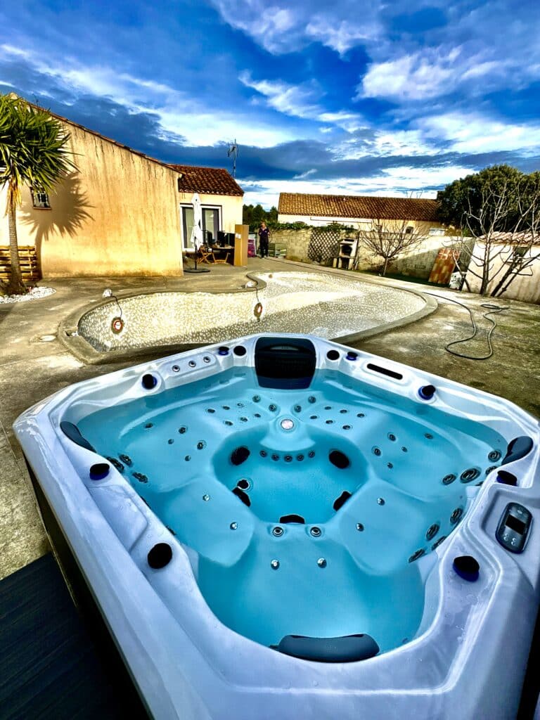 Spa moderne en extérieur avec eau turquoise vive, jets et panneau de contrôle. Ciel bleu intense sur patio et piscine décorative vide.