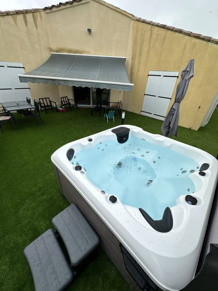 Jacuzzi moderne bouillonnant sur terrasse en gazon synthétique, avec mobilier de jardin et façade jaune sous un ciel de pluie.