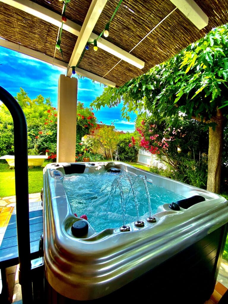 Jacuzzi moderne avec jets d'eau actifs et eau turquoise, abrité sous une pergola en chaume. Végétation luxuriante, fleurs rouges et ciel bleu intense.