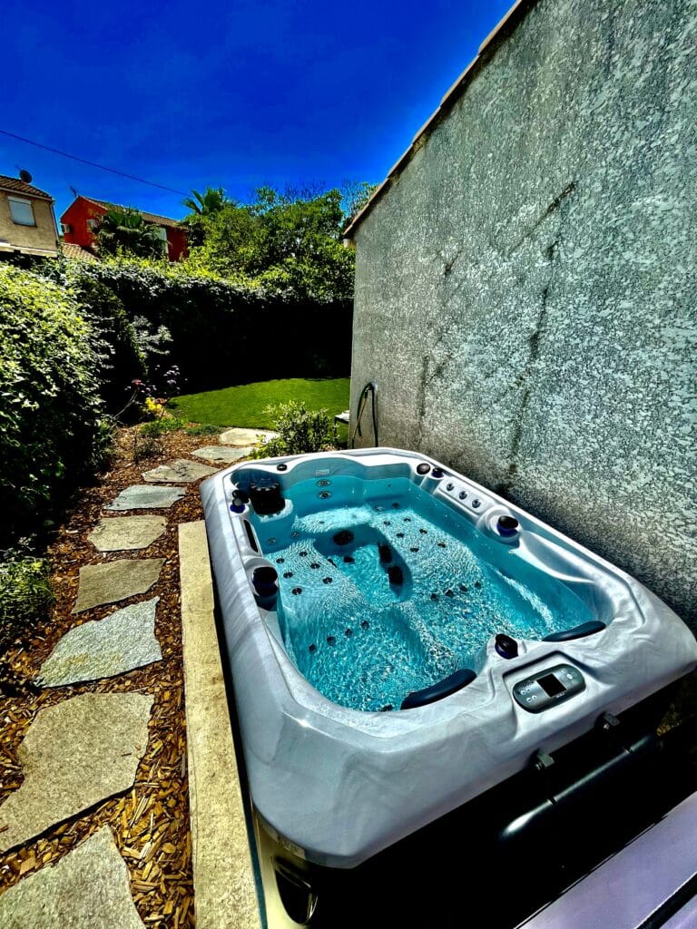 Jacuzzi jardin : luxe, eau turquoise et détente au soleil. Jacuzzi de jardin blanc aux jets activés, rempli d'eau turquoise. Allée de pierres et haie verte luxuriante sous un ciel bleu méditerranéen.