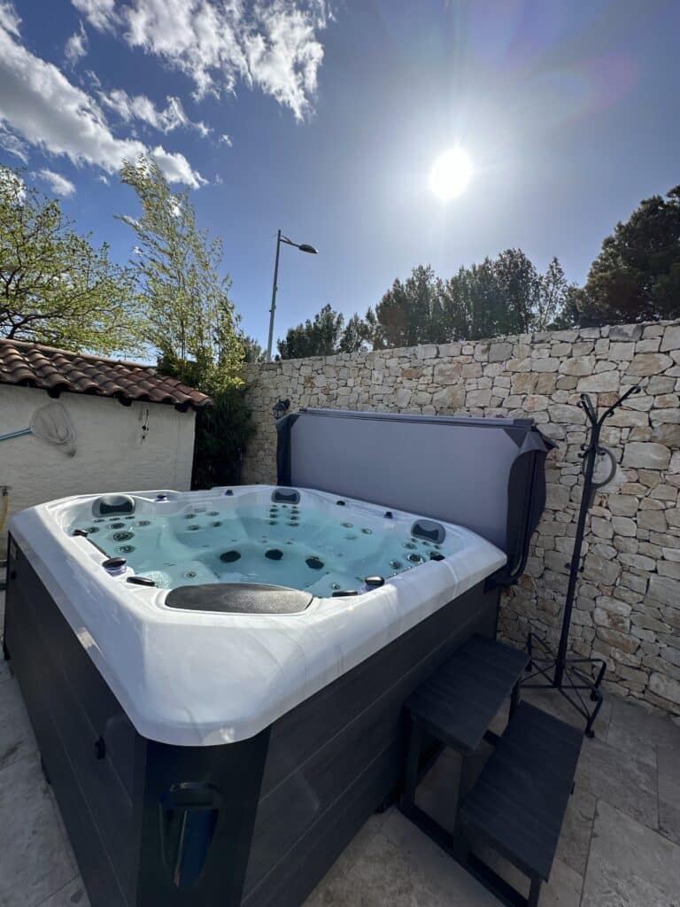 Jacuzzi extérieur moderne : détente sous le soleil Grand jacuzzi extérieur moderne blanc et gris foncé, rempli d'eau, installé contre un mur en pierres naturelles sous un ciel bleu éclatant.