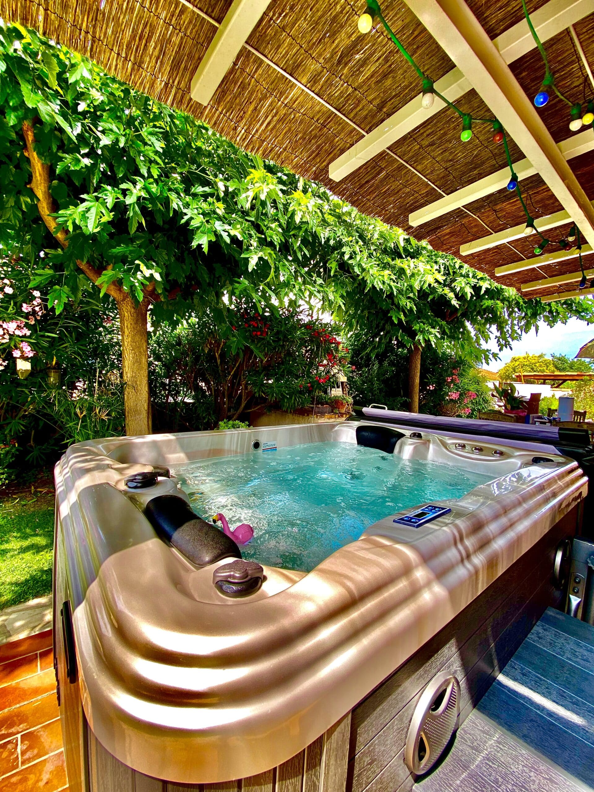 Jacuzzi extérieur turquoise sous paillote ombragée. Feuillage dense, guirlande lumineuse festive et petit flamant rose flottant.