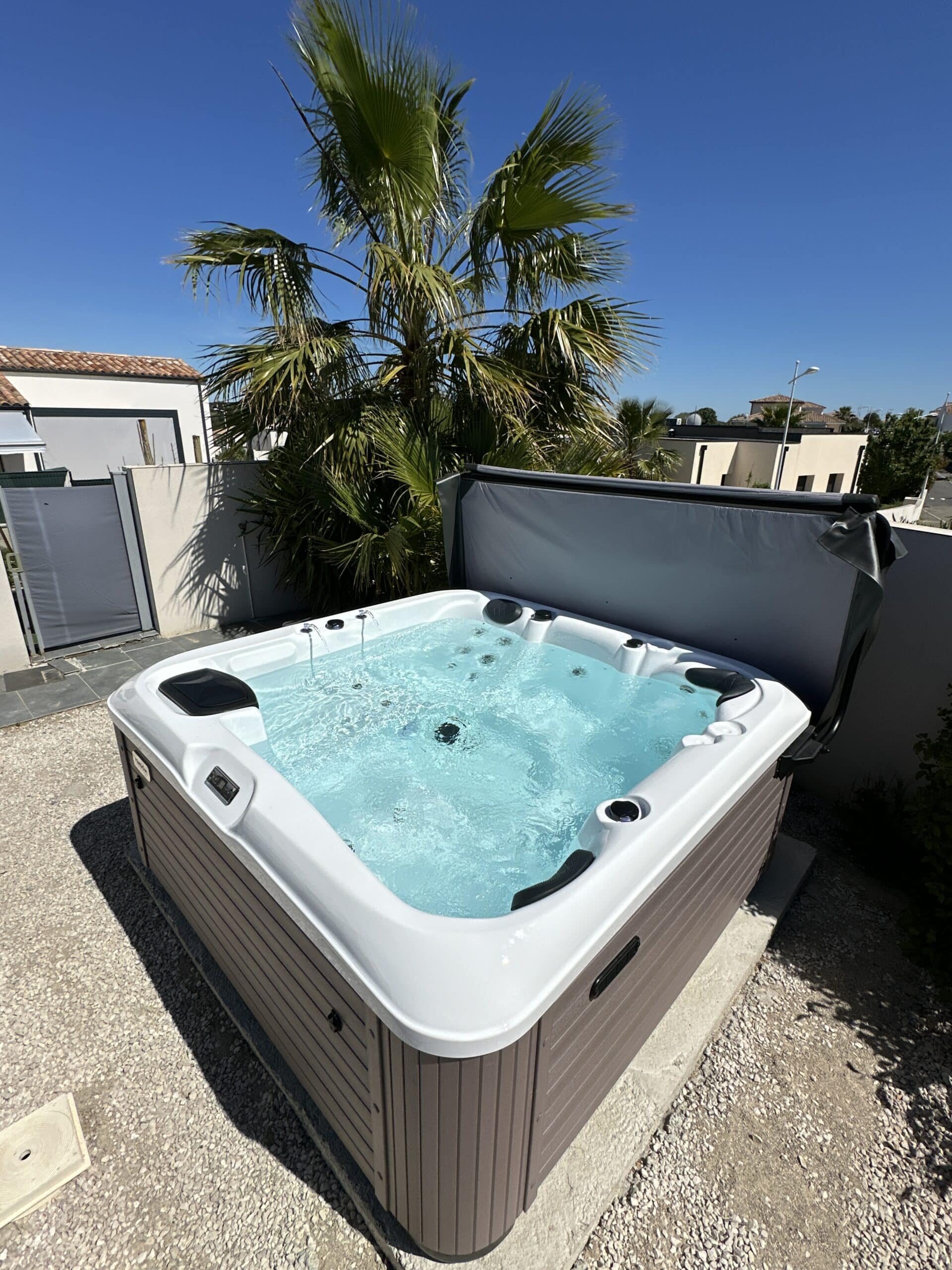Jacuzzi extérieur moderne avec eau turquoise bouillonnante, installé sur gravier, sous un grand palmier vert sur fond de ciel bleu intense.