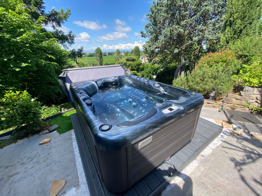 Jacuzzi extérieur luxe VENDOM avec vue panoramique. Luxueux spa VENDOM sur une terrasse en bois, eau turquoise. Vue panoramique ensoleillée sur la vallée et les arbres luxuriants.