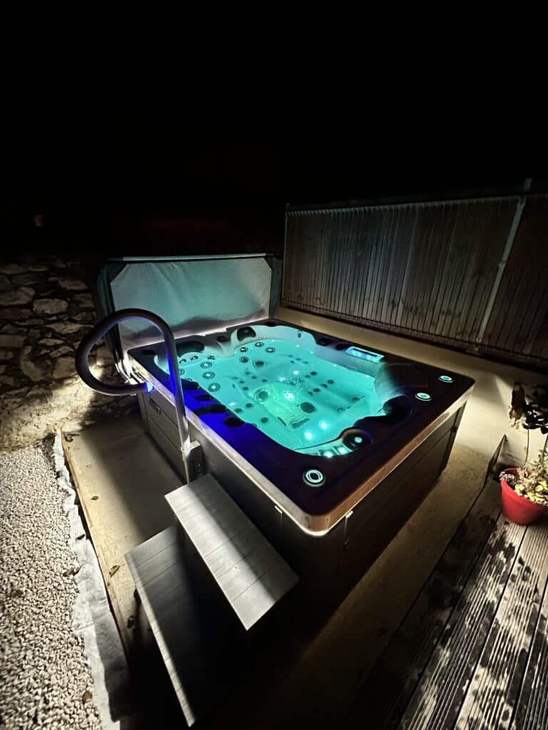 Jacuzzi extérieur de luxe éclairé en turquoise la nuit Luxueux spa d'extérieur la nuit, baigné d'une lumière turquoise vive. Entrée avec marches et rampe, entouré de pierres et de bois.