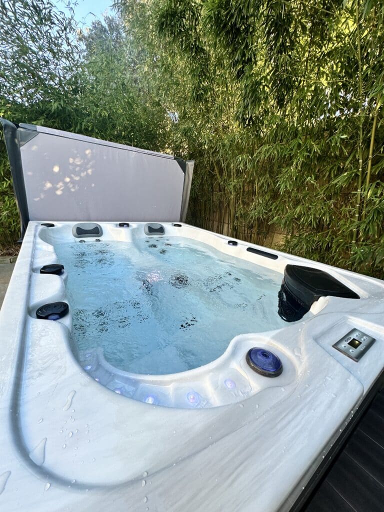 Jacuzzi extérieur au milieu des bambous : luxe et bien-être Spa extérieur blanc marbré, eau bouillonnante, couvercle gris relevé. Ambiance zen et luxuriante avec une haie de bambous verts en arrière-plan.